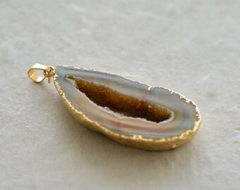 May include: A teardrop-shaped pendant made from a slice of agate with a gold-toned rim. The agate has a natural brown and white pattern with a hollow center.