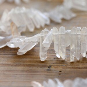 May include: A close-up of a strand of clear quartz crystals, arranged in a staggered pattern. The crystals are long and thin, with pointed ends. They are arranged on a wooden surface.