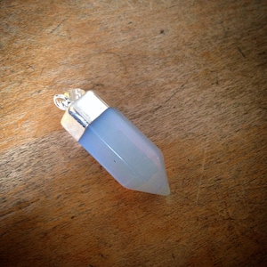 Puede incluir: Un colgante de cristal azul claro con una tapa y un lazo plateados. El cristal es de color azul pálido y tiene forma de prisma largo y puntiagudo. El colgante está sobre una superficie de madera.