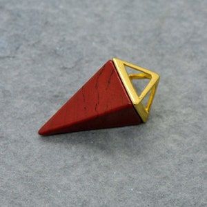 Op de afbeelding: Een rode edelsteen piramide hanger met een goudkleurig metalen frame. De hanger heeft een driehoekige vorm met een puntige punt.