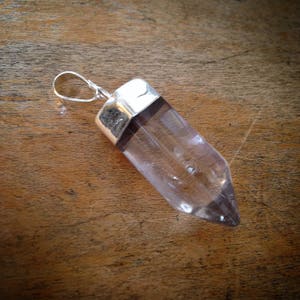 May include: A clear crystal point pendant with a silver setting. The pendant is shaped like a long, thin point and is suspended from a silver bail.