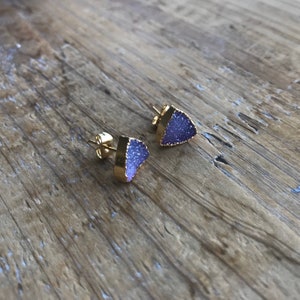 Puede incluir: Un par de pendientes de botón de oro con piedras triangulares de color púrpura. Las piedras tienen una textura brillante y brillante.