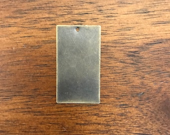 May include: A rectangular metal tag with a hole in the top. The tag is a dark bronze color and has a slightly textured surface.
