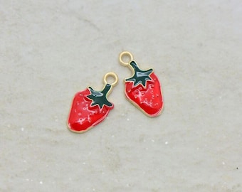Puede incluir: Dos colgantes de fresa roja con hojas verdes y detalles dorados. Los colgantes tienen un acabado brillante y un pequeño lazo en la parte superior para sujetarlos a joyas u otras manualidades. Miden aproximadamente 2 cm de largo.
