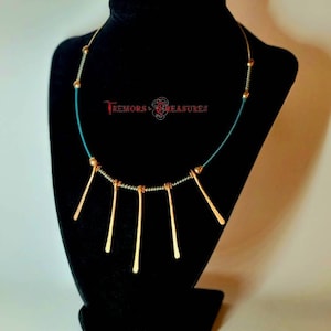 May include: A teal and gold necklace with a geometric design. The necklace has a series of thin, gold, vertical bars hanging from a teal cord with gold beads.
