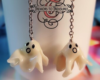 Op de afbeelding: Oorbellen in de vorm van geesten. Elke oorbel heeft een kleine, witte, spookvormige bedel met zwarte ogen en mond, die aan een zilveren ketting hangt. De tekst "Tremors to Treasures" is zichtbaar.