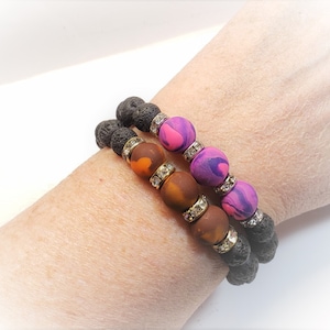 May include: Two beaded bracelets. One bracelet features lava rock beads, orange and brown beads, and gold-colored spacers with clear rhinestones. The other bracelet has lava rock beads, pink and purple beads, and gold-colored spacers with clear rhinestones.