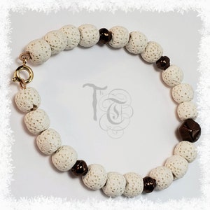 Może przedstawiać: Bransoletka z białych kamieni lawowych z brązowymi koralikami metalowymi. Bransoletka ma złote zapięcie.