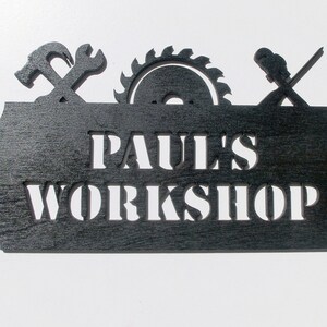 May include: Black wooden sign with the text "PAUL'S WORKSHOP" cut out. The sign is decorated with cut-out images of a hammer, wrench, and saw.