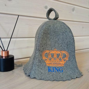 Peut inclure: Un chauffe-thé en feutre gris avec une poignée en boucle, brodé d'une couronne orange et du mot "KING" en bleu.