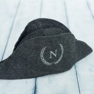 Puede incluir: Un sombrero bicornio de fieltro gris oscuro con una corona bordada blanca y la letra "N". El sombrero está doblado y descansa sobre una superficie de madera azul claro y blanca. El diseño recuerda la vestimenta militar histórica.