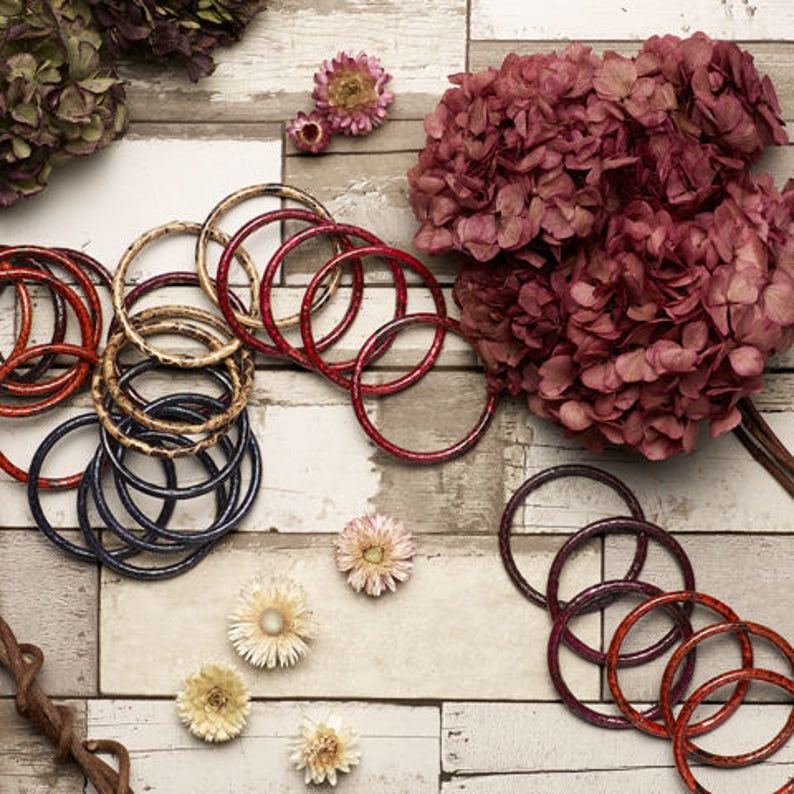 May include: A collection of colorful bangles in red, blue, and brown. The bangles are stacked and arranged on a white wooden surface. Dried flowers are scattered around the bangles.