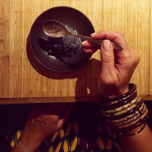 May include: A black bowl with a scoop of black ice cream on a bamboo mat. A hand is holding a spoon and scooping the ice cream.