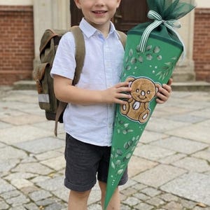 May include: A green cone-shaped school gift container featuring a brown teddy bear illustration and green leaf patterns. The top is finished with dark green tulle fabric and a striped ribbon bow. The item is held by a child wearing a white shirt.