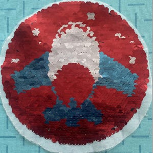 Può includere: Una toppa circolare con un design a paillettes caratterizzato da uno sfondo rosso, una forma centrale bianca e accenti blu. Le paillettes sono disposte per creare un motivo grafico su una base di tessuto azzurro con una trama a griglia visibile.