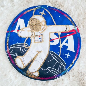 Könnte beinhalten: Ein runder blauer Patch mit einem weißen Astronauten in einem goldenen Raumanzug, umgeben von einem regenbogenfarbenen Rand. Der Patch trägt die Aufschrift "NASA" in weißen Buchstaben.