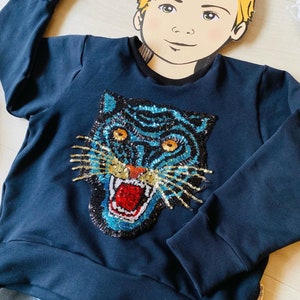 May include: Navy blue sweatshirt featuring a sequined tiger face design. The tiger has blue and gold sequins, with orange eyes and a red mouth. The sweatshirt has long sleeves and a crew neck.