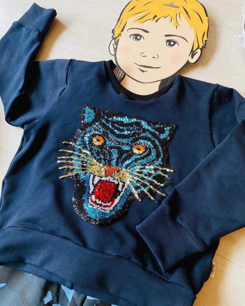 May include: Navy blue sweatshirt featuring a sequined tiger face design. The tiger has blue and gold sequins, with orange eyes and a red mouth. The sweatshirt has long sleeves and a crew neck.
