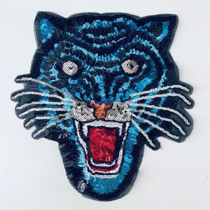 Könnte beinhalten: Ein Tigerkopf-Aufnäher aus Pailletten. Das Design hat ein Schwarz-Blau-Farbschema mit rotem Mund und oranger Nase. Dieser Aufnäher ist wahrscheinlich zum Verzieren von Kleidung oder Accessoires gedacht.