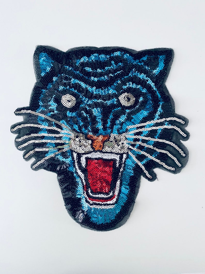 May include: A sequined tiger head patch with a fierce expression. The tiger features a black and blue color scheme with a red mouth and orange nose. The patch is likely for decorative purposes, such as embellishing clothing or accessories.