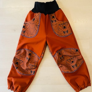 Może przedstawiać: Rdzawobrązowe spodnie z czarną gumką w pasie i wzorem niedźwiedzi w okularach. Spodnie mają kieszenie po bokach i łaty na kolanach.