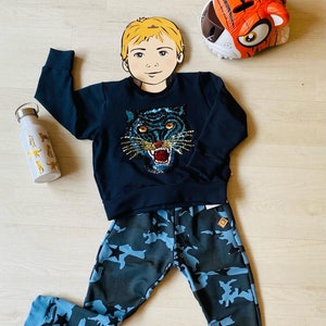 May include: A navy blue sweatshirt with a sequined tiger face design, paired with camouflage print pants featuring blue stars. A white water bottle with animal illustrations and a tiger-themed headpiece complete the ensemble.