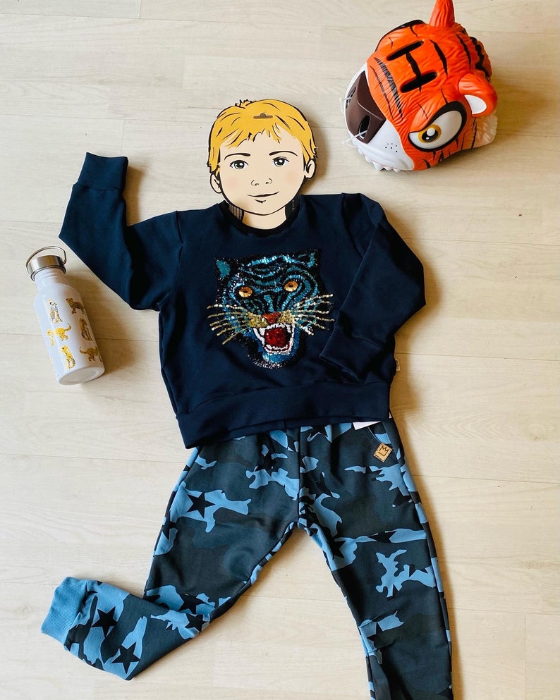 May include: A navy blue sweatshirt with a sequined tiger face design, paired with camouflage print pants featuring blue stars. A white water bottle with animal illustrations and a tiger-themed headpiece complete the ensemble.