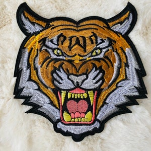 Può includere: Un patch ricamato a forma di testa di tigre. La tigre ha un'espressione feroce con i denti scoperti e gli occhi gialli. Il patch è di colore arancione e giallo vibrante con dettagli in nero e bianco.