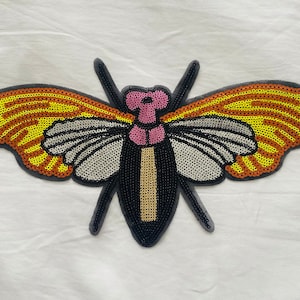 May include: A black, yellow, and orange sequined butterfly applique with pink accents. The butterfly has a detailed design and is perfect for adding a touch of whimsy to clothing or crafts.