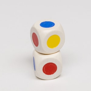 Puede incluir: Dos dados de madera blancos con puntos de colores. El dado de arriba tiene un punto azul, un punto rojo y un punto amarillo. El dado de abajo tiene un punto azul y un punto rojo.