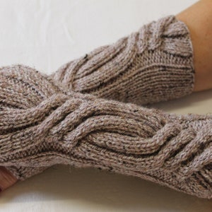 Puede incluir: Un par de guantes sin dedos de punto en un color marrón neutro moteado. Los guantes presentan un diseño de punto de cable en la parte superior y están diseñados para cubrir la mano y la muñeca.