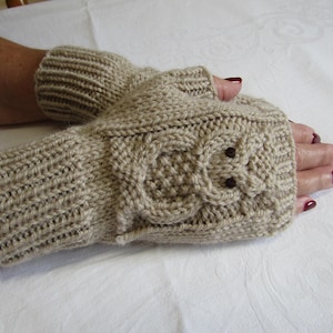 May include: Beige knitted fingerless gloves with an owl design. The gloves are made of a soft, textured yarn and feature a detailed owl face with two small brown button eyes. The wrist cuffs are ribbed.