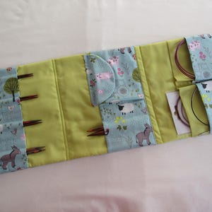 May include: A light green and blue fabric knitting needle case with a farm animal pattern. The case is open, revealing knitting needles and circular needles. The case is approximately 30 cm wide and 20 cm tall.