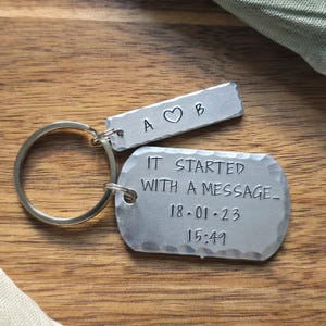 May include: A silver-toned metal keyring featuring two rectangular tags with hammered edges. The smaller tag is stamped with A, a heart symbol, and B. The larger tag is stamped with the text IT STARTED WITH A MESSAGE, 18.01.23, and 15:49.