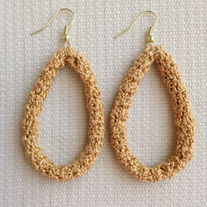 May include: A pair of gold crochet earrings with a teardrop shape. The earrings are made with a beige yarn and have a subtle shimmer.
