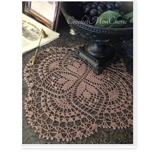 May include: A brown crocheted doily with a delicate floral design. The doily is placed on a dark countertop with a black bowl of grapes and a framed picture in the background.