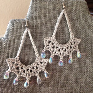 May include: A pair of crocheted earrings with iridescent teardrop beads hanging from the bottom. The earrings are a light beige color and have a delicate, lacy design.
