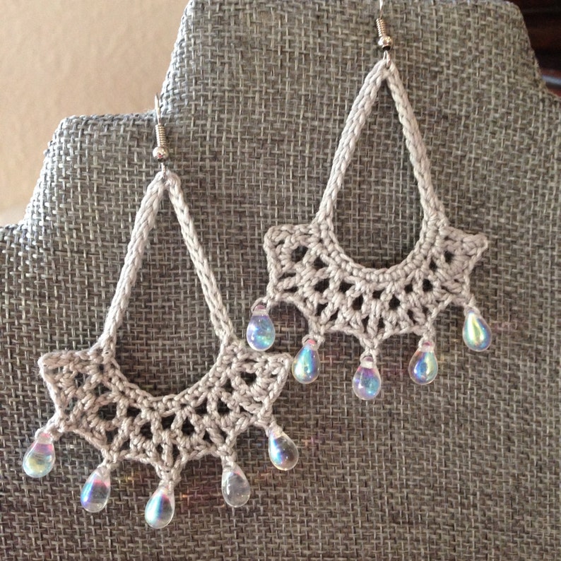 May include: A pair of crocheted earrings with iridescent teardrop beads hanging from the bottom. The earrings are a light beige color and have a delicate, lacy design.