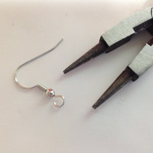 May include: A silver earring hook with a small ball at the end. The hook is open and ready to be attached to an earring. A pair of silver pliers are also in the image.