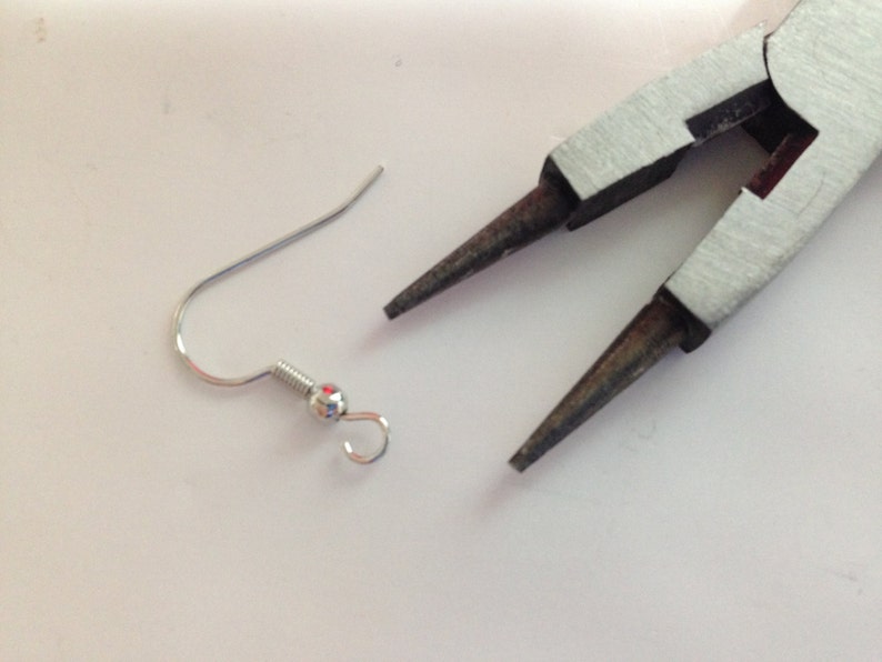 May include: A silver earring hook with a small ball at the end. The hook is open and ready to be attached to an earring. A pair of silver pliers are also in the image.