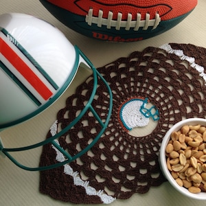 May include: A white football helmet with a teal and orange stripe design, a brown crocheted doily with a white center, and a bowl of peanuts.