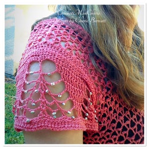 May include: Close-up of a pink crocheted top with a delicate lace pattern and silver beads. The top features a unique openwork design on the sleeves.