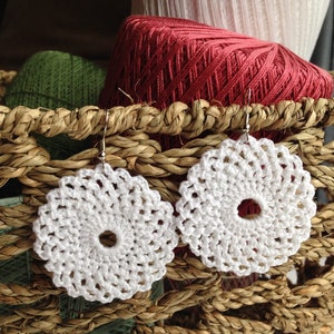 May include: A pair of white crocheted earrings with a circular design. The earrings are hanging from a wicker basket filled with yarn.