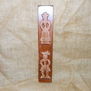 Puede incluir: Un molde de galletas de madera vintage con dos figuras talladas: una mujer con un vestido y un hombre con calzones y sombrero. Las figuras son de color claro sobre la madera marrón oscura. El molde es rectangular.