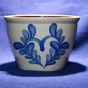 May include: A ceramic planter with a cream-colored base and a blue floral design. The planter has a wide opening and a slightly tapered shape. The blue design is symmetrical and features stylized leaves. The interior is brown.
