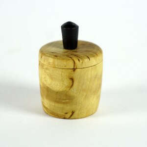 May include: A small, round wooden box with a black knob on the lid. The box is made of light wood with a natural grain pattern.