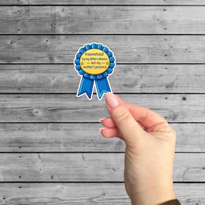 May include: A blue and yellow ribbon sticker with the text "traumatized by my father's absence - and my mother's presence." The sticker is held by a hand against a gray wooden background.