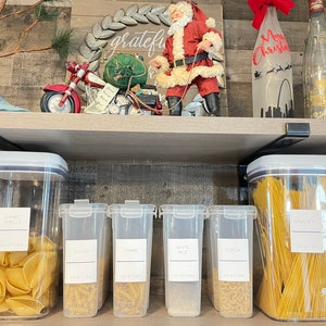May include: Six clear plastic containers with labels on a wooden shelf. The containers hold various dry goods, including pasta, rice, and spaghetti. The labels are white with black text and say "Jumbo Shells", "Rotini", "Penne", "White Rice", "Elbow", and "Spaghetti".