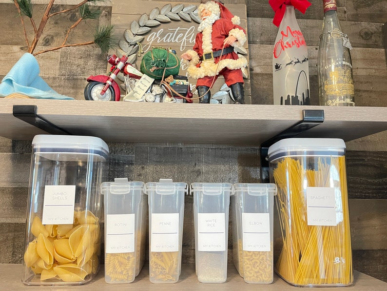 May include: Six clear plastic containers with labels on a wooden shelf. The containers hold various dry goods, including pasta, rice, and spaghetti. The labels are white with black text and say "Jumbo Shells", "Rotini", "Penne", "White Rice", "Elbow", and "Spaghetti".