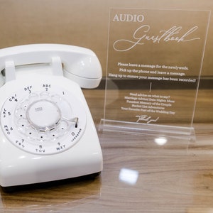 May include: A white rotary dial phone with a white coiled cord. The phone is on a wooden surface with a sign that says "Audio Guestbook". The sign has instructions for leaving a message for the newlyweds.